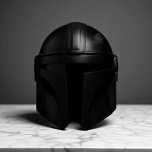 May include: A matte black Mandalorian helmet with a sleek, angular design. The helmet features a T-shaped visor and a crest with the letters "SN". The helmet is displayed on a white marble surface.