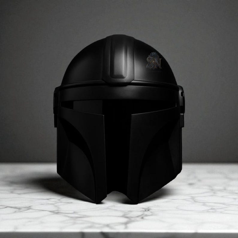 May include: A matte black Mandalorian helmet with a sleek, angular design. The helmet features a T-shaped visor and a crest with the letters "SN". The helmet is displayed on a white marble surface.