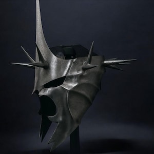 May include: A metallic gray helmet with a menacing design. The helmet features a tall central spike and multiple smaller spikes around the edges. The face area has a stylized, open design. The overall aesthetic suggests a fantasy or historical theme.