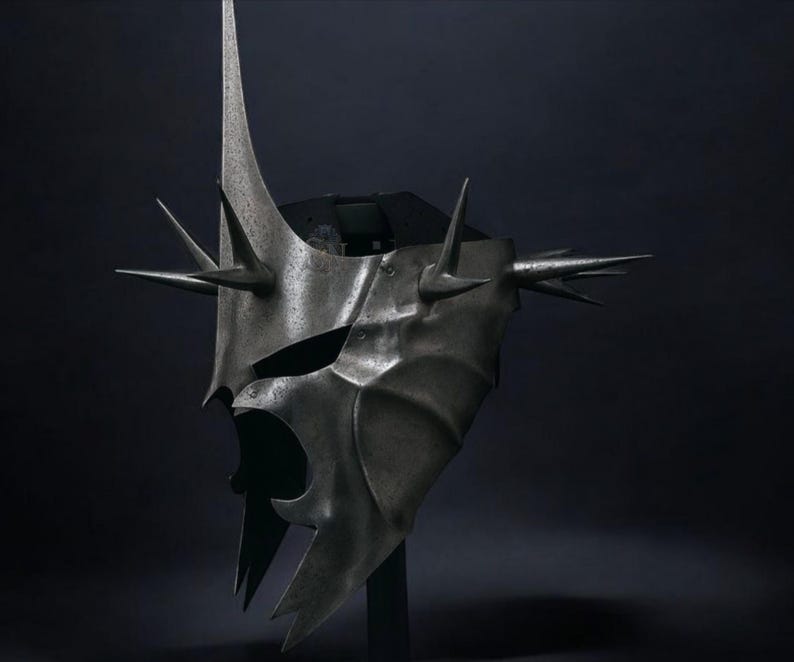 May include: A metallic gray helmet with a menacing design. The helmet features a tall central spike and multiple smaller spikes around the edges. The face area has a stylized, open design. The overall aesthetic suggests a fantasy or historical theme.