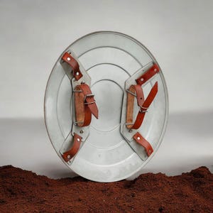 Puede incluir: Un escudo plateado con correas y hebillas de cuero marr&oacute;n. El escudo tiene un dise&ntilde;o circular con anillos conc&eacute;ntricos. Las correas de cuero est&aacute;n remachadas al escudo.