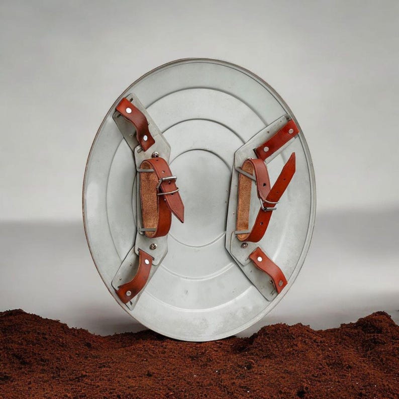 Puede incluir: Un escudo plateado con correas y hebillas de cuero marr&oacute;n. El escudo tiene un dise&ntilde;o circular con anillos conc&eacute;ntricos. Las correas de cuero est&aacute;n remachadas al escudo.
