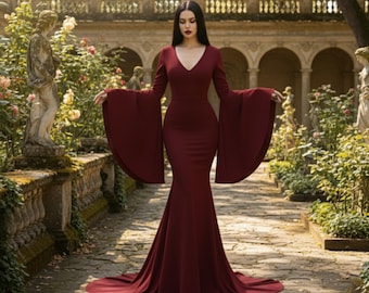 May include: A long, burgundy dress with a V-neck and dramatic bell sleeves. The dress is form-fitting through the bodice and flares out into a flowing skirt. The woman is standing in a garden setting with stone statues and a stone walkway.