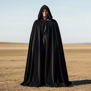 May include: A full-length black hooded cloak made of a soft, flowing fabric. The cloak features a deep hood and a tie closure at the neck. The person is standing in a field with a clear sky in the background.