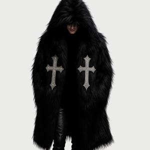 May include: A long, black faux fur coat with a large hood. The coat features two silver-colored crosses on the front. The coat is worn over a black turtleneck and black pants.