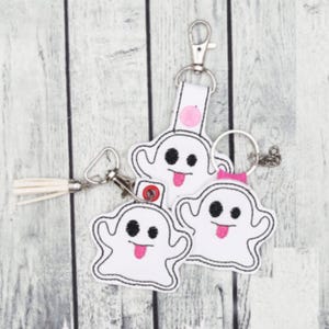 May include: White ghost-shaped keyrings with black eyes, pink tongues, and black outlines. One keyring has a pink circle and a white strap. Another has a pink bow. A white tassel is attached to one keyring.