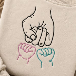 May include: A beige sweatshirt featuring an embroidered design of three hands in a fist bump formation. The top hand is outlined in black thread, while the two smaller hands below are outlined in pink and teal thread respectively.