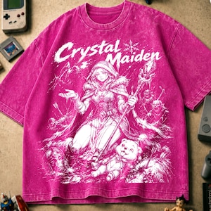 Puede incluir: Una camiseta magenta con efecto lavado al ácido que presenta un estampado gráfico blanco de una figura encapuchada sosteniendo un bastón con el texto Crystal Maiden. El diseño incluye una pequeña criatura, flechas y elementos de fantasía.