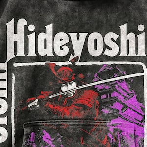 Puede incluir: Sudadera negra con la palabra "Hideyoshi" en blanco. Debajo, un gráfico representa un samurái con armadura roja sosteniendo una espada, con un edificio morado al fondo. El diseño tiene un aspecto vintage.