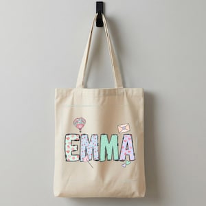 May include: A cream-coloured canvas tote bag featuring the name "EMMA" in colourful letters. The letters are decorated with hearts, a hot air balloon, an envelope, and other small graphics. White trainers are below the bag.