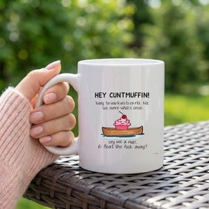 May include: White ceramic mug with the text "HEY CUNTMUFFIN!" and other text. A cartoon image of a cupcake in a canoe is also on the mug. The mug has a curved handle and is sitting on a dark surface.