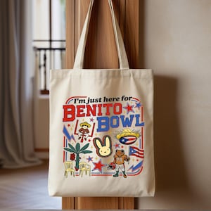 May include: Beige tote bag with the text "I'm just here for BENITO BOWL" in red and blue. The bag features colorful illustrations of a rabbit, frog, stars, and other graphics. The bag has two beige handles.
