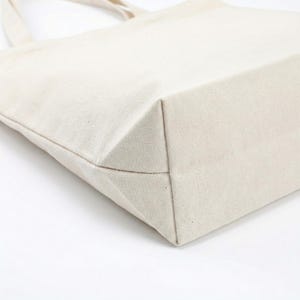 Puede incluir: Primer plano de un bolso tote de lona beige. El bolso tiene una base rectangular y un dise&ntilde;o sencillo. La tela parece ser un material tejido resistente. Las asas del bolso son visibles en el fondo.