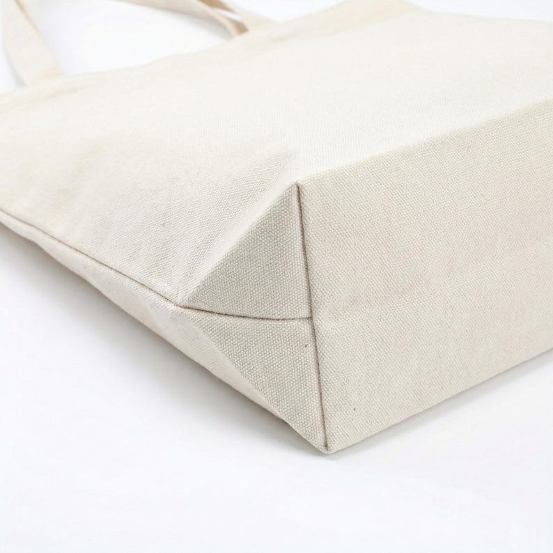 Puede incluir: Primer plano de un bolso tote de lona beige. El bolso tiene una base rectangular y un dise&ntilde;o sencillo. La tela parece ser un material tejido resistente. Las asas del bolso son visibles en el fondo.