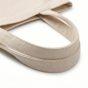 Puede incluir: Primer plano de un bolso tote de lona beige. La imagen destaca las asas resistentes, con doble costura, y la tela natural sin blanquear. El dise&ntilde;o del bolso sugiere que es adecuado para llevar comestibles, libros u otros art&iacute;culos cotidianos.