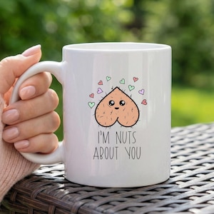 Pode incluir: Caneca de cerâmica branca com um design humorístico. A caneca apresenta uma ilustração de desenho animado de uma noz com um rosto e pequenos corações. O texto "I'M NUTS ABOUT YOU" é impresso abaixo. A caneca é segurada por uma pessoa.