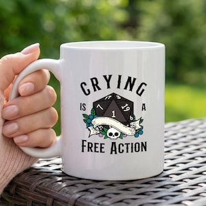 Puede incluir: Taza de cerámica blanca con el texto "Crying is a Free Action" en letras negras. La taza presenta un gráfico de dados negro con detalles florales y de calaveras. La taza está sostenida por una persona.