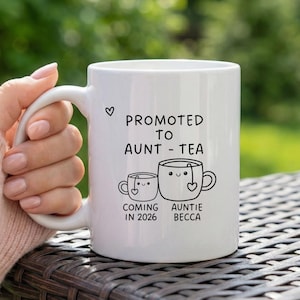 Puede incluir: Taza de cerámica blanca con el texto "PROMOTED TO AUNT - TEA" y dos tazas de té de dibujos animados. Una taza dice "COMING IN 2026" y la otra dice "AUNTIE BECCA". Un pequeño corazón está dibujado encima del texto.