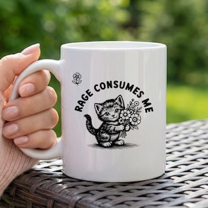 May include: White ceramic mug with a black and white kitten illustration holding flowers, and the text "Rage Consumes Me". The mug has a curved handle and a floral design near the top.