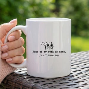May include: White ceramic mug with a black cat illustration and the text "None of my work is done. put I sure an." The mug has a curved handle and is held by a person.