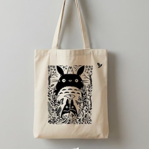 Puede incluir: Bolso tote beige con una ilustración en blanco y negro de dos personajes de Totoro rodeados de mariposas y follaje. El diseño incluye un Totoro grande y uno más pequeño, con un fondo de bosque. El bolso tiene dos asas.