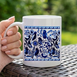 May include: White ceramic mug featuring a blue and white bird and floral pattern. The design includes various bird illustrations, flowers, and foliage. The mug is held by a person's hand, with a blurred green background.