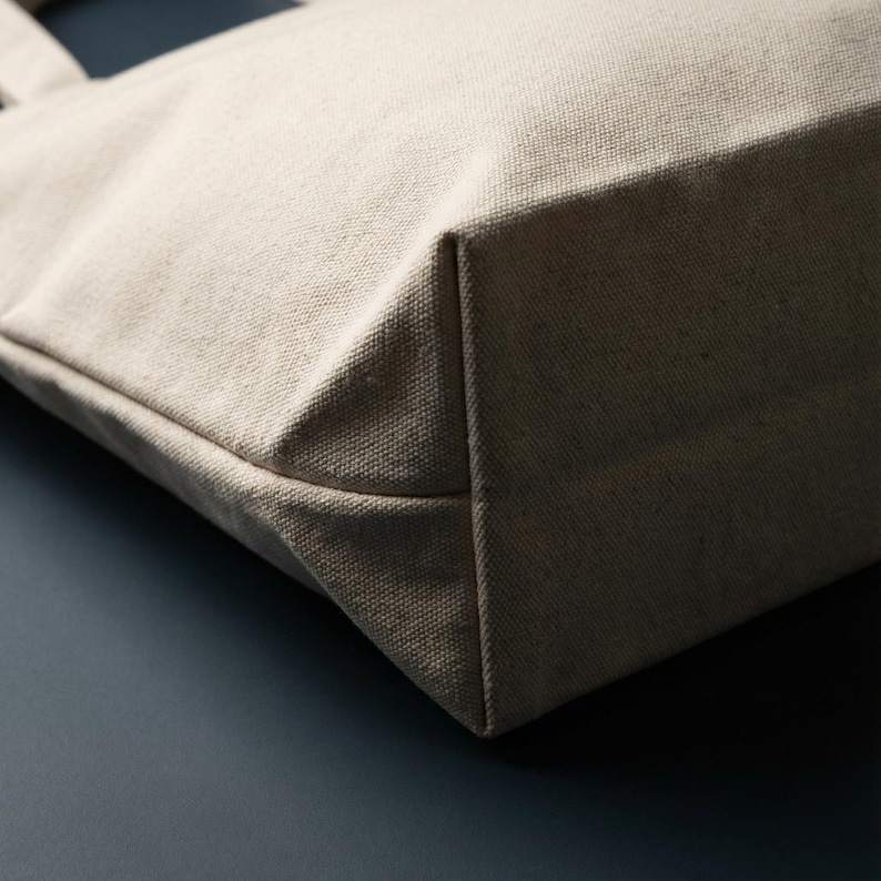 Puede incluir: Primer plano de un bolso tote de lona beige. El bolso tiene una base rectangular y un dise&ntilde;o simple y minimalista. La tela parece ser un material tejido resistente, y el bolso probablemente est&aacute; dise&ntilde;ado para transportar objetos.