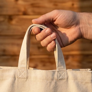 Puede incluir: Una mano sostiene una bolsa de lona beige con asas resistentes. La tela de la bolsa tiene un aspecto texturizado y las asas est&aacute;n bien sujetas. El fondo es una superficie de madera.