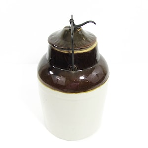 May include: A brown and white glazed ceramic jar with a metal lid and handle. The jar has a white base and a brown top.