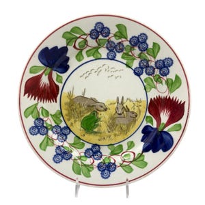May include: A white ceramic plate with a hand-painted design. The plate features a central scene of rabbits and a frog in a field, surrounded by floral and berry motifs in blue, red, and green. A red border frames the edge.
