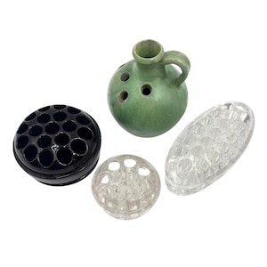 May include: Four flower frogs in various shapes and colors. A black, round frog, a clear, oval frog, a clear, round frog, and a green, jug-shaped frog with a handle and holes for flower stems.