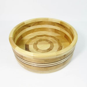 May include: A round wooden bowl with a light and dark wood grain pattern. The bowl has a circular design on the base and is approximately 10 inches in diameter. The bowl's exterior features two dark bands. The bowl is made of various wood pieces.