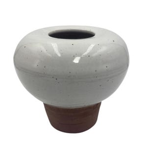 May include: A white ceramic vase with a rounded body and a dark opening at the top. The vase has a speckled texture and sits on a brown, cylindrical base. This decorative vase is suitable for displaying flowers or as a standalone art piece.