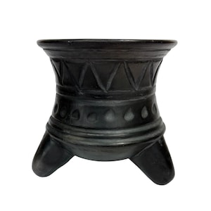May include: A dark, handcrafted, three-legged vessel with a wide, open top. The vessel features carved geometric patterns, including triangles and teardrop shapes, around its body. The overall design suggests a rustic, artisanal aesthetic.