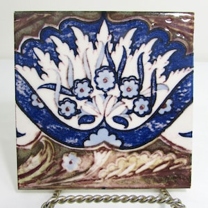 May include: A square ceramic tile with a blue and white floral design. The tile has a brown border and a white background. The floral design is intricate and detailed, with leaves and flowers in a repeating pattern.
