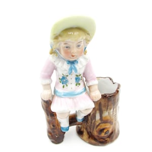 May include: A vintage porcelain figurine of a young girl in a pink dress and a yellow hat, perched on a brown tree stump. The girl's dress is adorned with blue floral details and a blue ribbon.