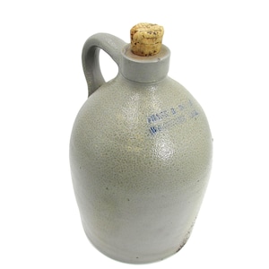 May include: A gray stoneware jug with a handle and a cork stopper. The jug has a rounded body and a textured surface. Blue text is visible on the front of the jug. The jug is likely used for storing liquids.