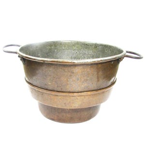 May include: A vintage copper basin with two curved handles. The basin has a wide, open top and a tiered design, with a slightly smaller base. The copper has a weathered, aged appearance, with variations in color and patina.