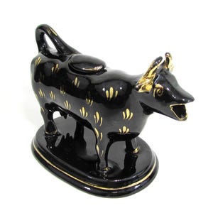 May include: Black ceramic cow creamer with gold accents. The cow-shaped creamer has a handle, a spout, and is decorated with gold leaf patterns. The creamer sits on an oval base with a gold trim. A decorative kitchenware item.
