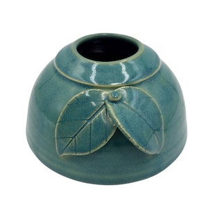 May include: A teal ceramic vase with a rounded body and a wide opening. Two stylized leaves are attached to the front of the vase. The vase has a glossy finish and a dark interior.