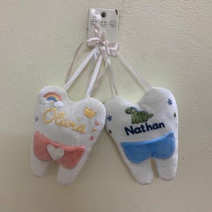 May include: Two white plush tooth-shaped cushions hanging from a white wall hook. One cushion features the name Olivia in yellow, a rainbow, a crown, and a pink pocket with a white heart. The other features the name Nathan in blue and a green dinosaur.