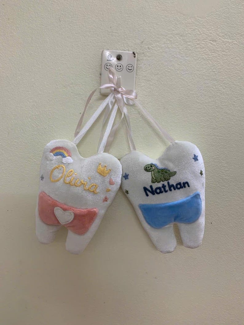 May include: Two white plush tooth-shaped cushions hanging from a white wall hook. One cushion features the name Olivia in yellow, a rainbow, a crown, and a pink pocket with a white heart. The other features the name Nathan in blue and a green dinosaur.
