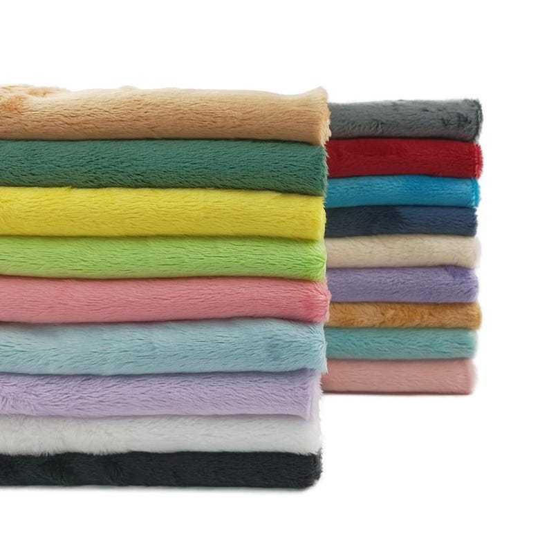 K&ouml;nnte beinhalten: Gestapelte, farbenfrohe Pl&uuml;schstoffe in verschiedenen Farbt&ouml;nen, darunter Beige, Gr&uuml;n, Gelb, Rosa, Blau, Lila, Wei&szlig; und Schwarz. Das weiche, strukturierte Material ist ordentlich gefaltet und zeigt eine Reihe von Farbt&ouml;nen. Das Bild ist gut beleuchtet und hebt die Pl&uuml;schstruktur hervor.