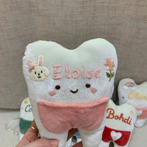 May include: A white plush tooth-shaped pillow featuring an embroidered face, a pink pocket, and the name Eloise in pink thread. The pillow is decorated with an embroidered bunny and a flower. A white ribbon is attached to the bottom of the pillow.