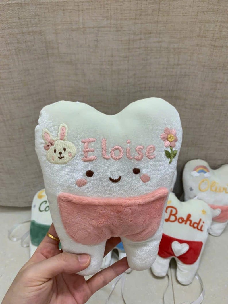 May include: A white plush tooth-shaped pillow featuring an embroidered face, a pink pocket, and the name Eloise in pink thread. The pillow is decorated with an embroidered bunny and a flower. A white ribbon is attached to the bottom of the pillow.