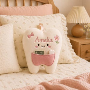 May include: A cream-coloured plush tooth pillow featuring a pink pocket, embroidered with the name Amelia in pink script. The tooth has a smiling face, a small pink bow, and a pink flower. A small white heart and a banknote are inside the pocket.