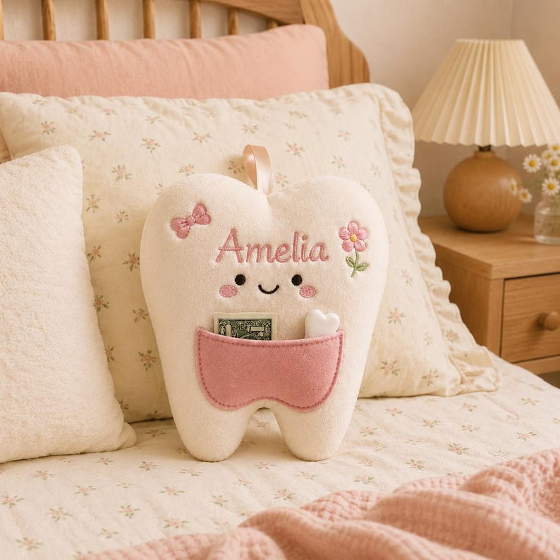 May include: A cream-coloured plush tooth pillow featuring a pink pocket, embroidered with the name Amelia in pink script. The tooth has a smiling face, a small pink bow, and a pink flower. A small white heart and a banknote are inside the pocket.