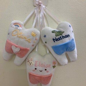 May include: Three white plush tooth-shaped pillows with pockets, each hanging by a white ribbon. One features the name Olivia with a rainbow, one says Nathan with a green dinosaur, and one displays Eloise with a bunny face and pink flower.
