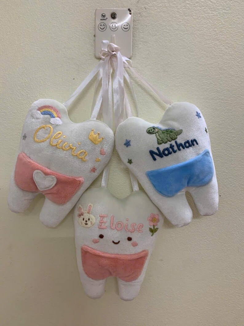 May include: Three white plush tooth-shaped pillows with pockets, each hanging by a white ribbon. One features the name Olivia with a rainbow, one says Nathan with a green dinosaur, and one displays Eloise with a bunny face and pink flower.