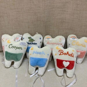 May include: A collection of plush tooth-shaped pillows in off-white fabric, each featuring embroidered names including Cooper, Nathan, Noah, Annemarie, Bohdi, and Olivia. The pillows have small colourful pockets and attached white ribbons.